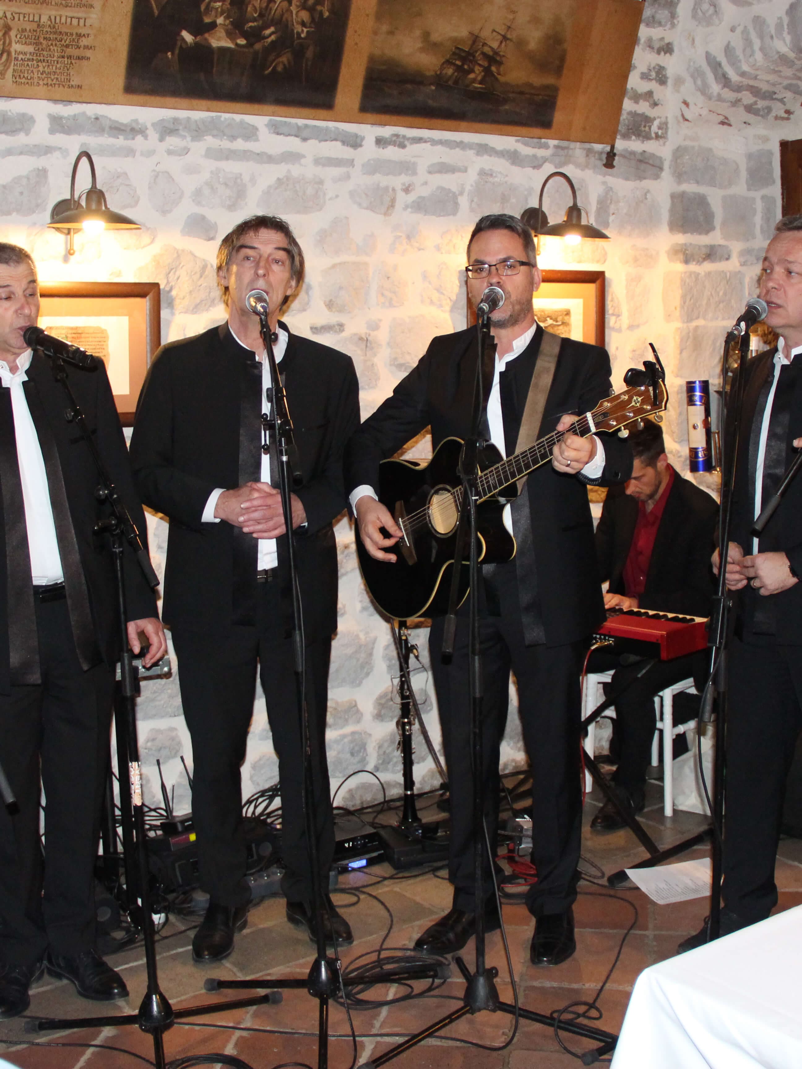 Musicians performing live at Restaurant Conte, with a guitarist and singers in black suits, against a stone wall.