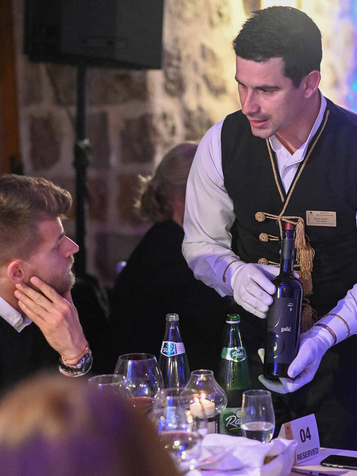 A server presenting a bottle of wine to a guest at restaurant Conte, with a stone wall backdrop and elegantly set dining table