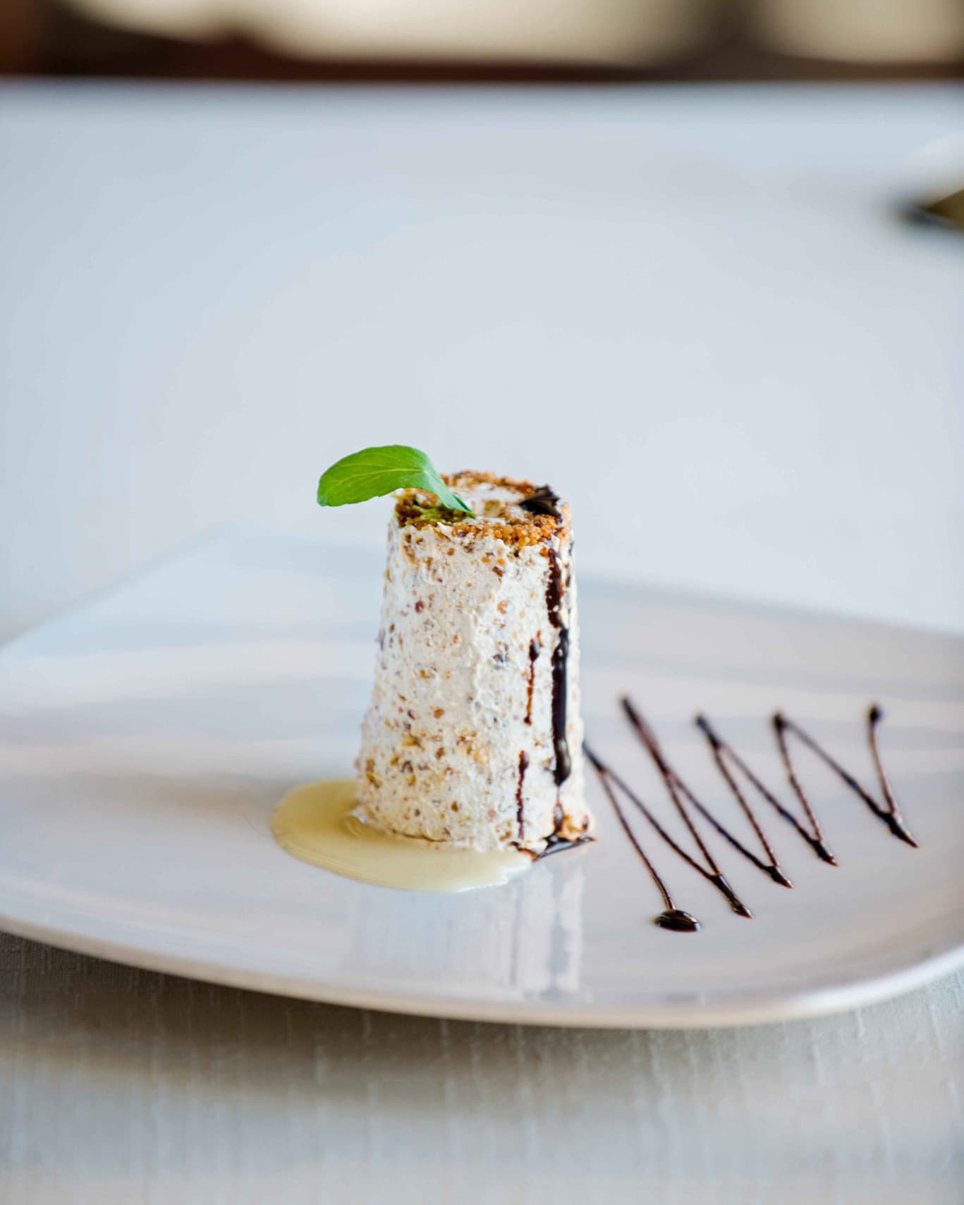 Exquisite dessert at Hotel Conte's restaurant, featuring a cylindrical pastry garnished with a mint leaf and drizzled with chocolate sauce on a white plate.