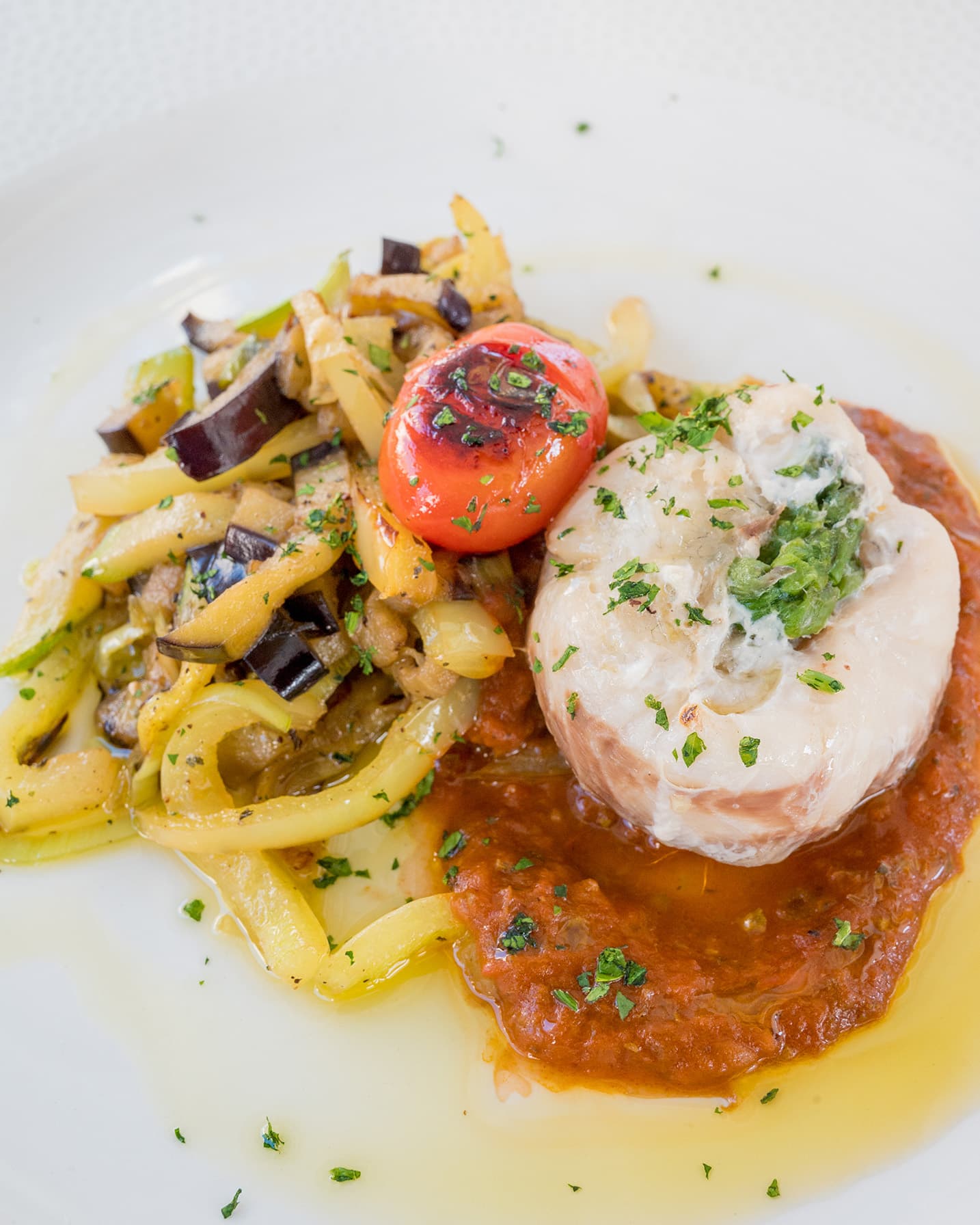 Gourmet dish at Hotel Conte's restaurant featuring a fillet of fish topped with herbs, accompanied by sautéed vegetables and a cherry tomato on a bed of tomato sauce.