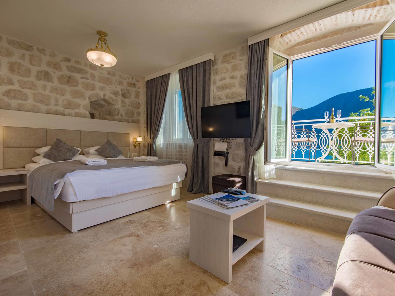 Luxurious hotel room at Hotel Conte featuring a comfortable bed, stone walls, a flat-screen TV, and a balcony with a view of the Bay of Kotor.