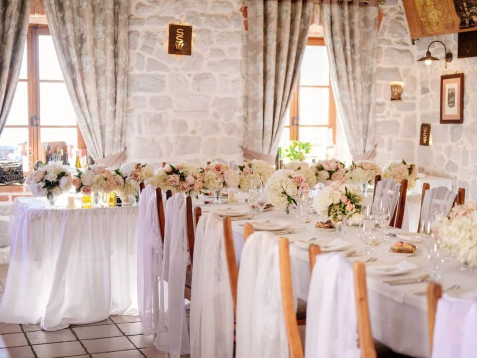 Beautiful indoor wedding reception setup at Hotel Conte, featuring tables decorated with white tablecloths, blush and white floral arrangements, and elegant place settings against stone walls and large windows.