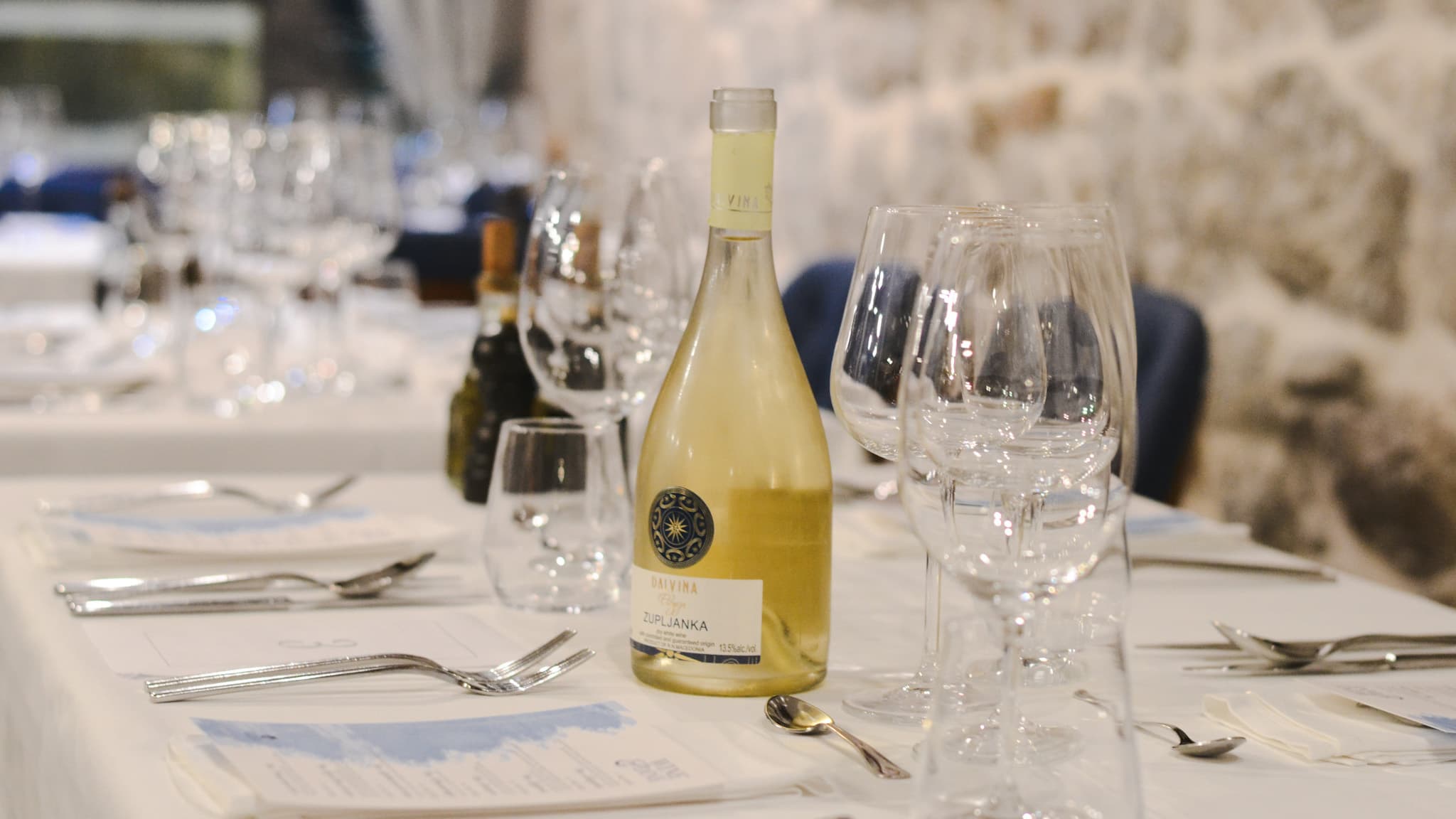 Elegant table setting with a bottle of white wine, wine glasses, and cutlery in a rustic stone-walled venue.