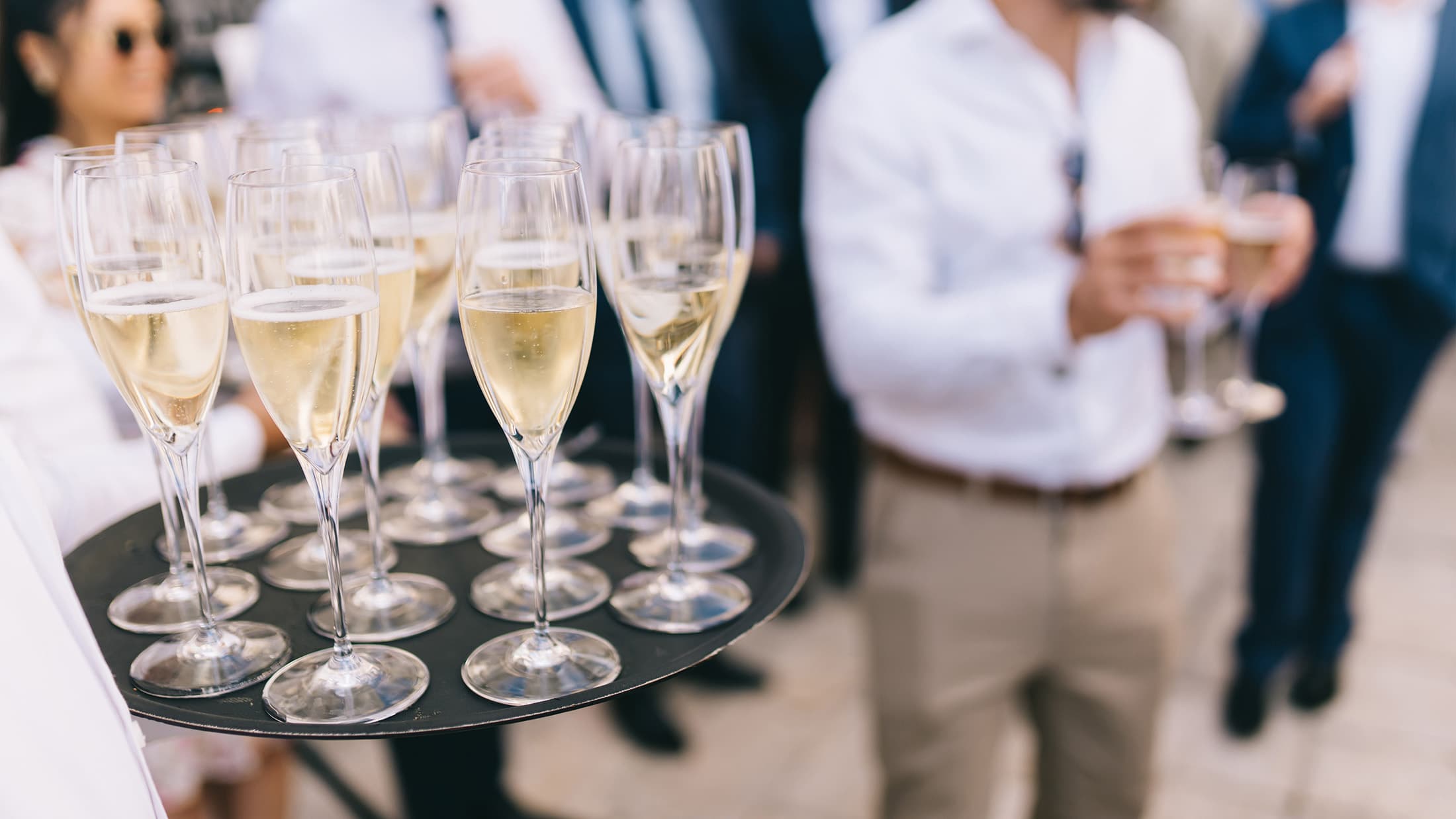 Waiter holding a tray with wine glasses, with guests in the background.