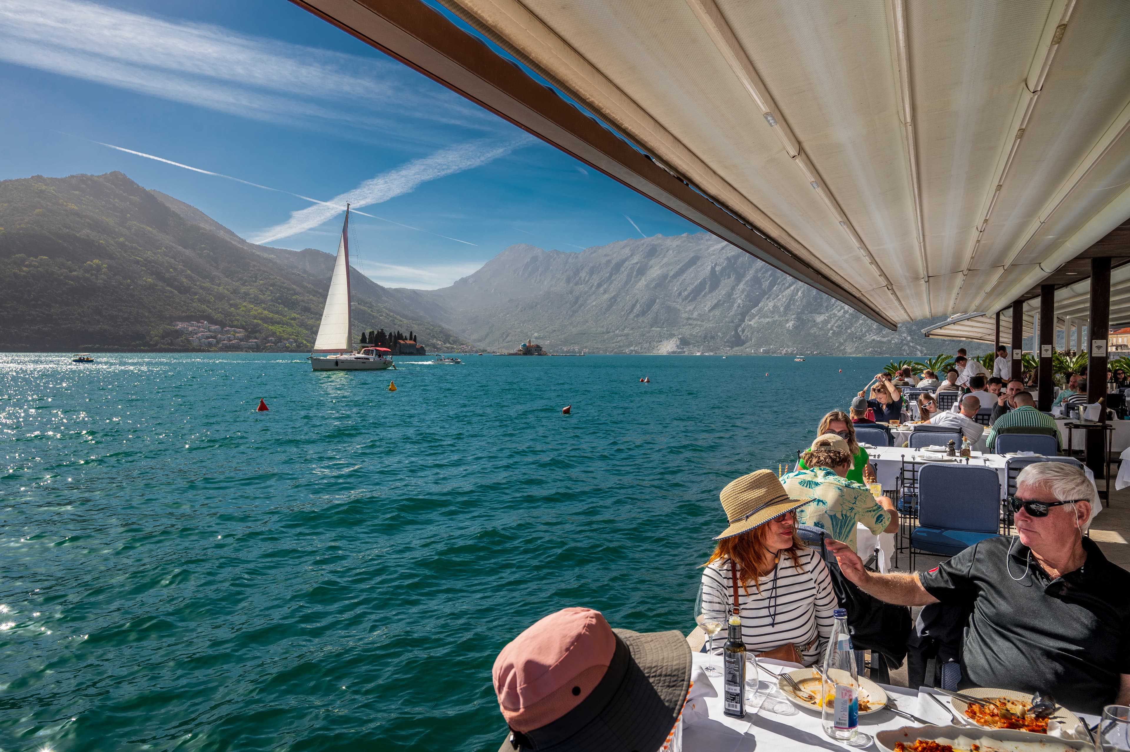 Guests dining on the terrace of Hotel Conte's restaurant with stunning views of the Bay of Kotor, a sailboat, and surrounding mountains.