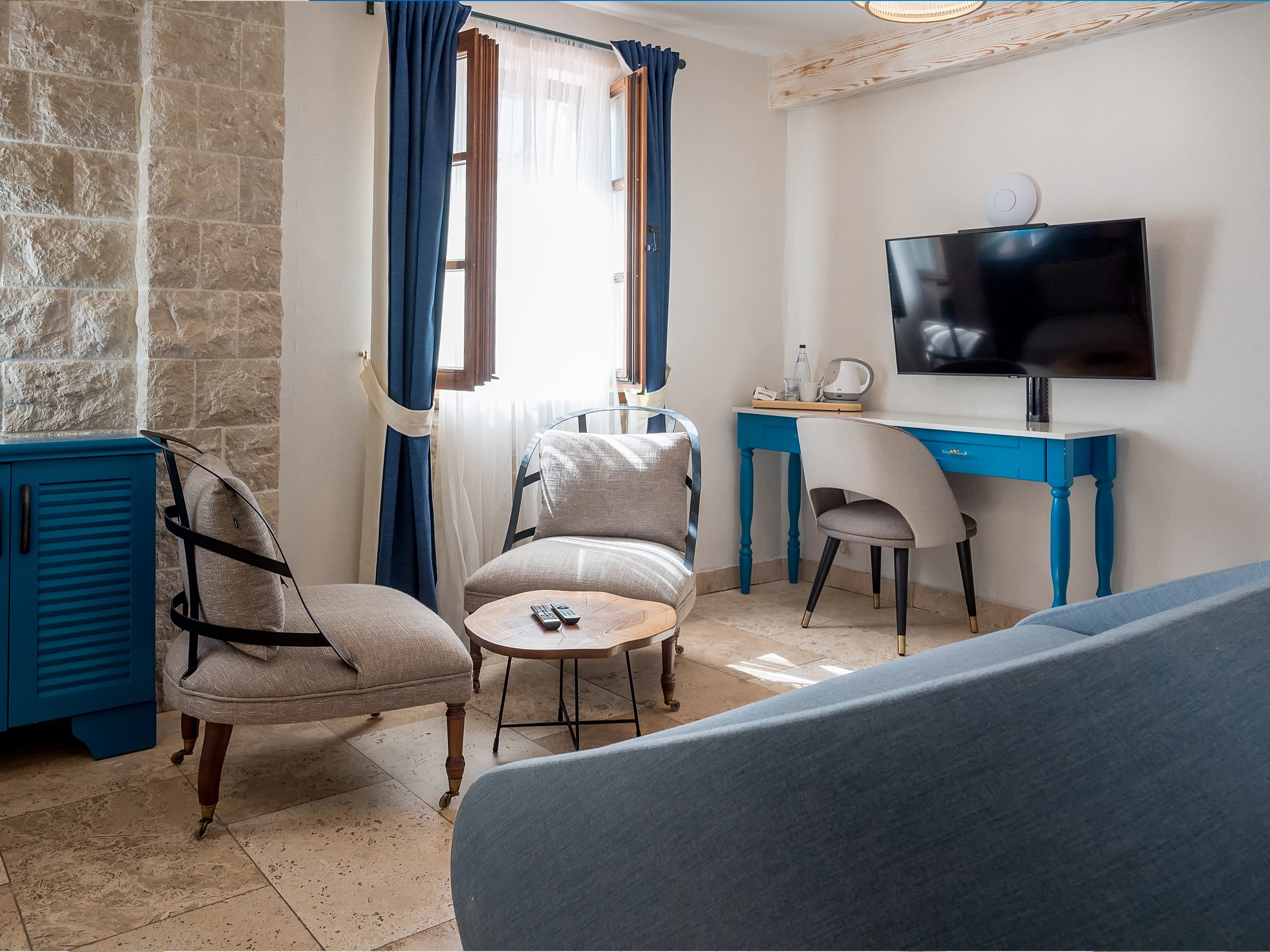 Cozy sitting area in a hotel room at Hotel Conte featuring comfortable chairs, a blue desk, a flat-screen TV, and natural stone walls.