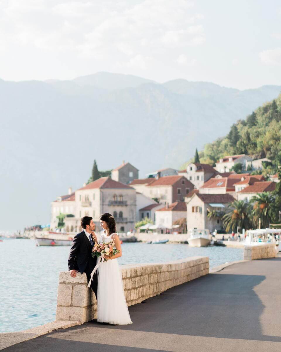 Bride and groom sharing a romantic moment on the waterfront in Perast, with charming historic buildings and mountains in the background, near Hotel Conte.