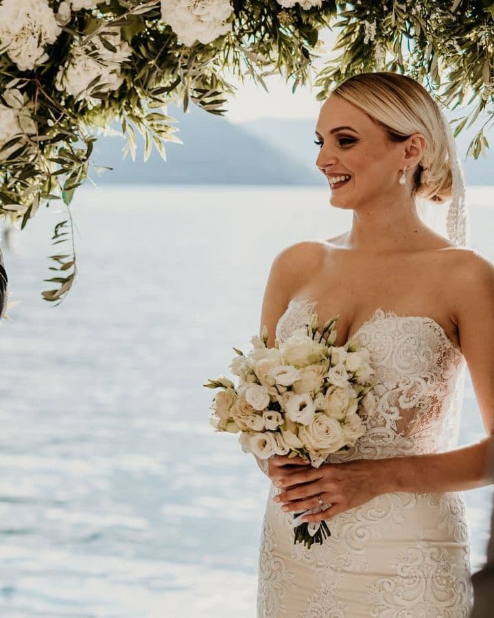 Smiling bride holding a bouquet of white flowers, standing under a floral arch with the Bay of Kotor in the background at a wedding at Hotel Conte.