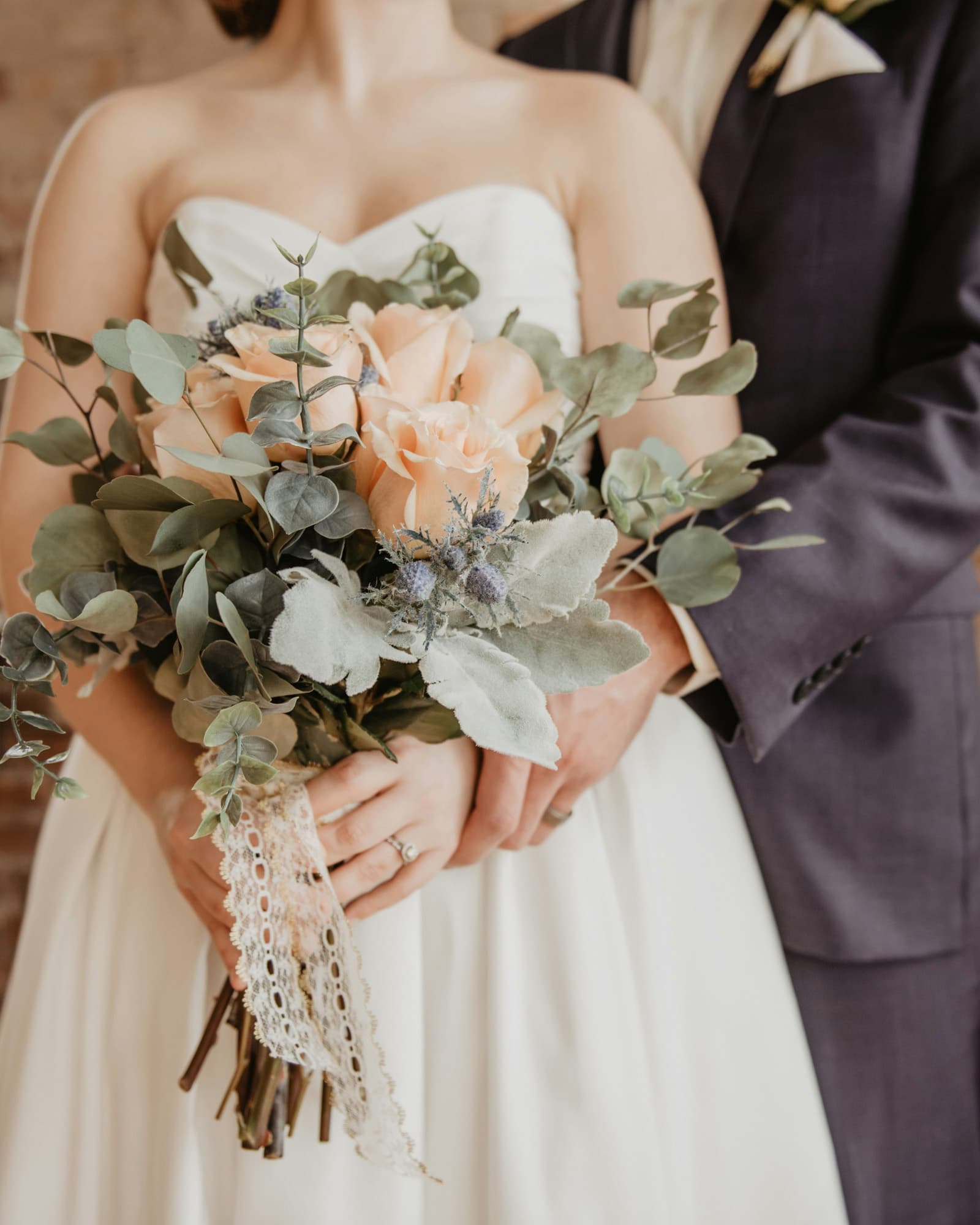 Bride holding a bouquet of peach roses and greenery, with the groom standing behind her.