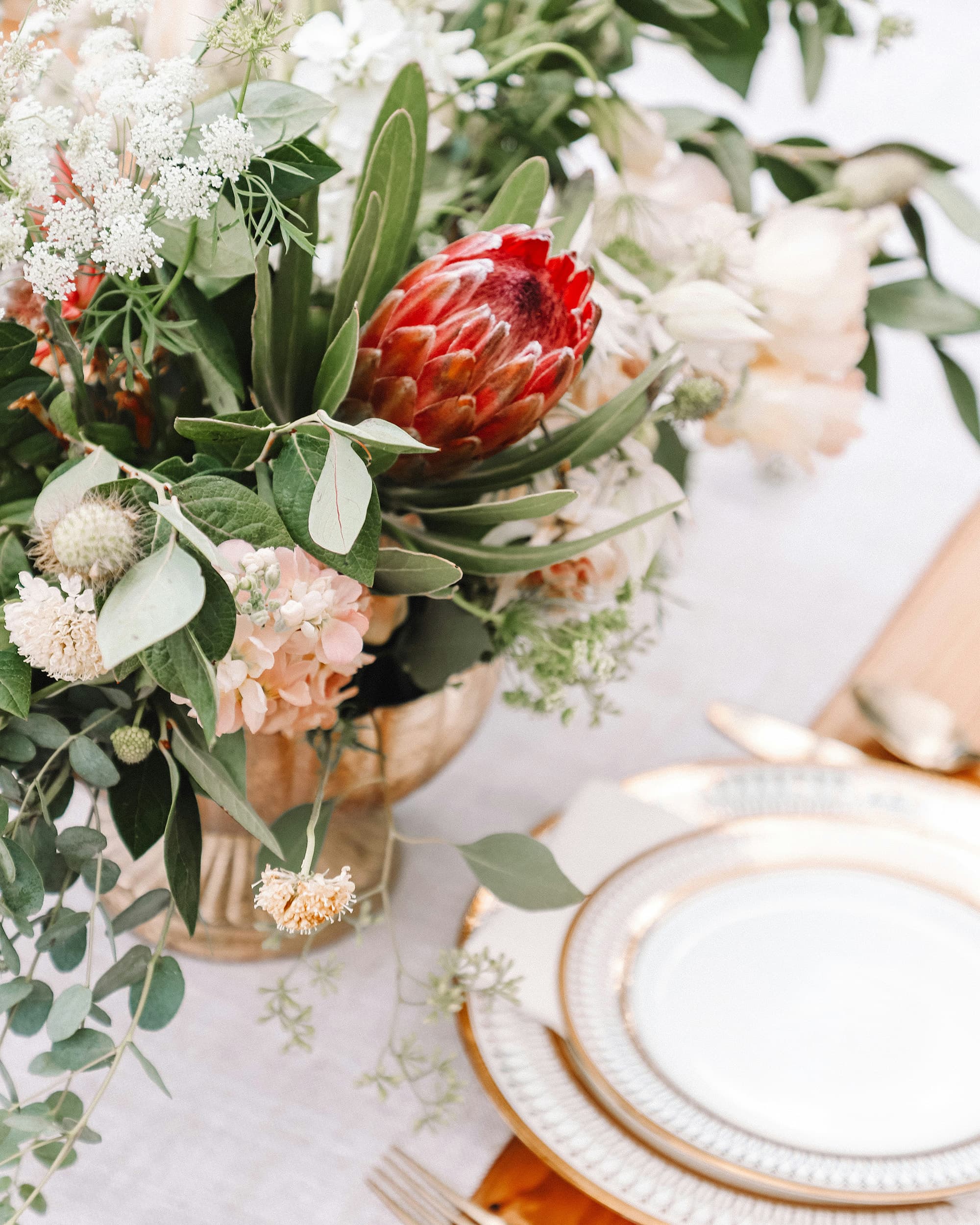 Elegant floral centerpiece with a mix of white and blush flowers, greenery, and a prominent red protea, set on a table with fine china.