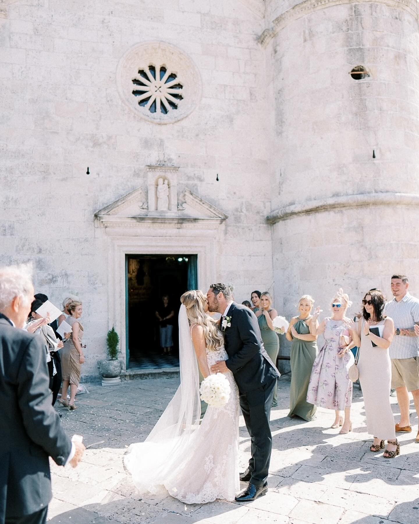 Bride and groom sharing a kiss outside a historic church in Perast, surrounded by celebrating guests