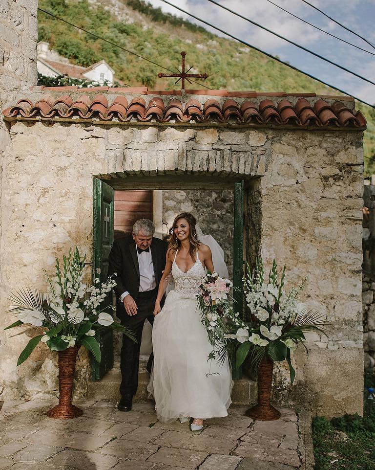 Bride walking out of a historic stone chapel with her father, surrounded by floral arrangements and greenery at Hotel Conte.