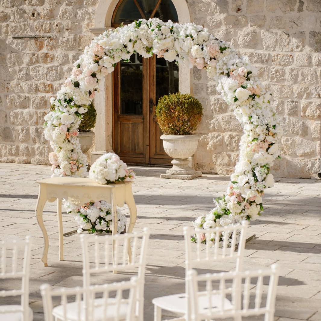 Wedding ceremony setup at Hotel Conte, featuring a floral arch decorated with white and blush flowers, white chairs, and a small table, set against a stone wall backdrop.