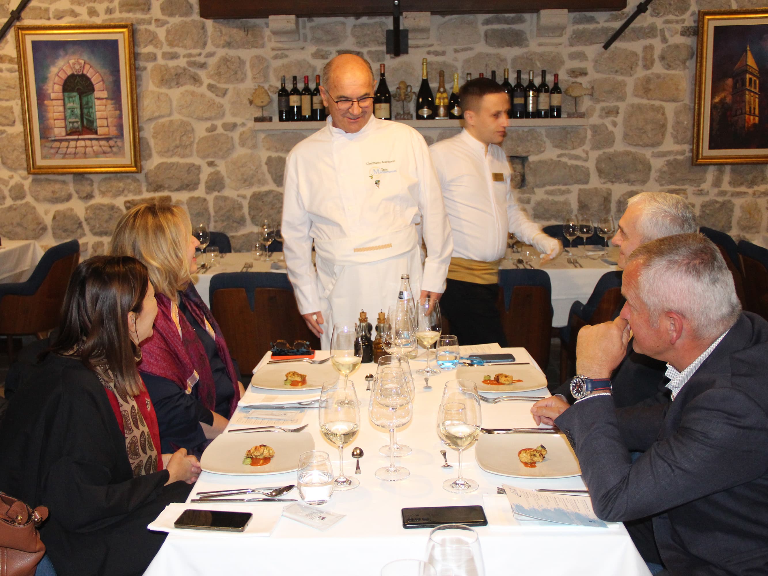 Chef conversing with guests at a dining table in Restaurant Conte, with plates of gourmet food and wine glasses.